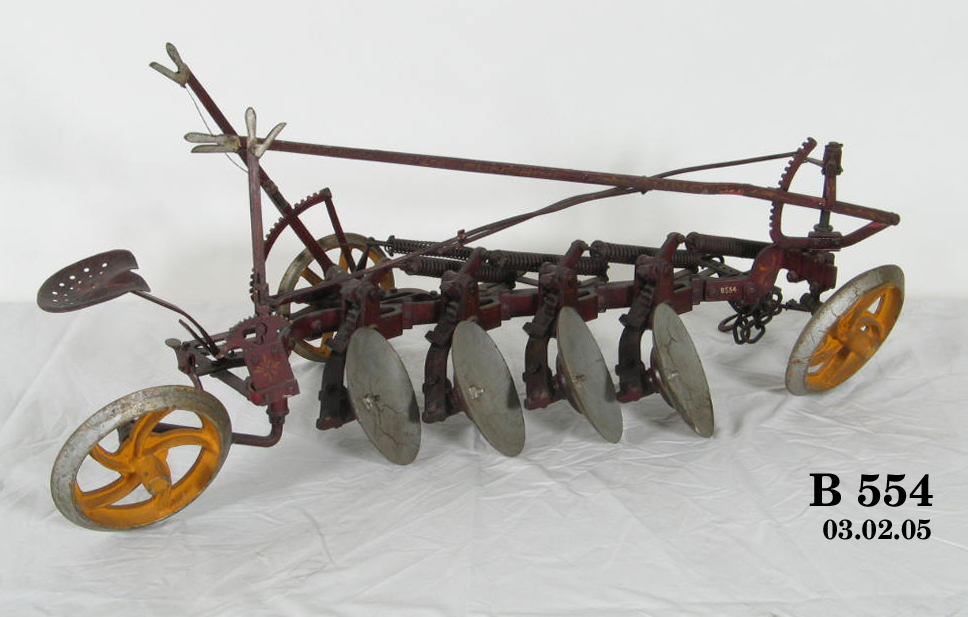 Model of a type of plough with four metal discs for turning the soil, a seat for the operator and three handles to make adjustments, all set on a frame with three wheels. It is finished in red and yellow.